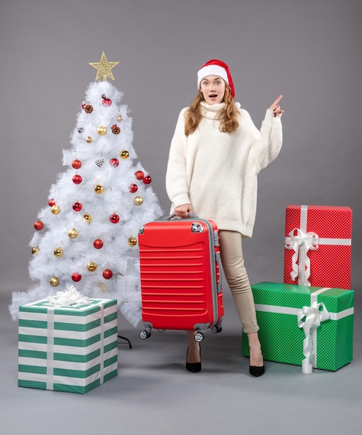 La vista frontale ha sorpreso la ragazza di natale con il cappello della santa che tiene la sua borsa da viaggio rossa vicino all'albero di natale bianco