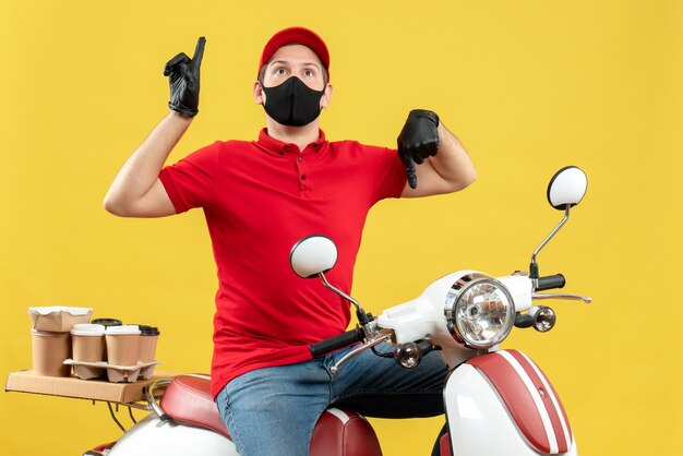 Front view of surprised young adult wearing red blouse and hat gloves in medical mask delivering order sitting on scooter with thumbs up and down on yellow background