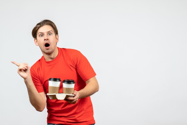 Front view of surprised young guy in red blouse holding coffee in paper cups and pointing something on the right side on white background