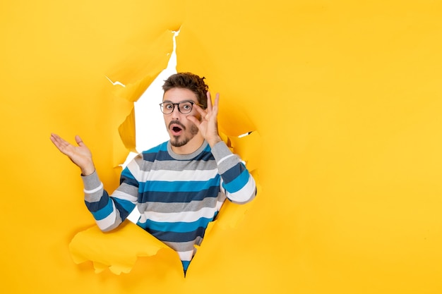 Front view surprised young man holding his eyeglasses peeping through hole in paper yellow wall