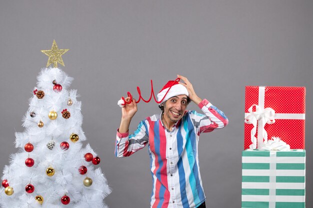 Вид спереди удивил молодого человека, держащего свою спиральную весеннюю шляпу Санта возле белой рождественской елки