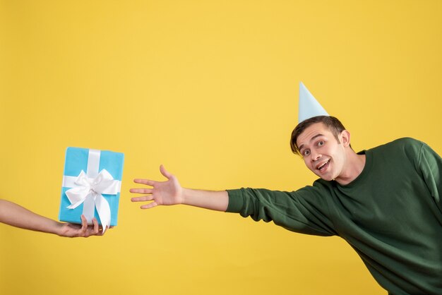 Front view surprised young man with party cap trying to catch gift in human hand on yellow 