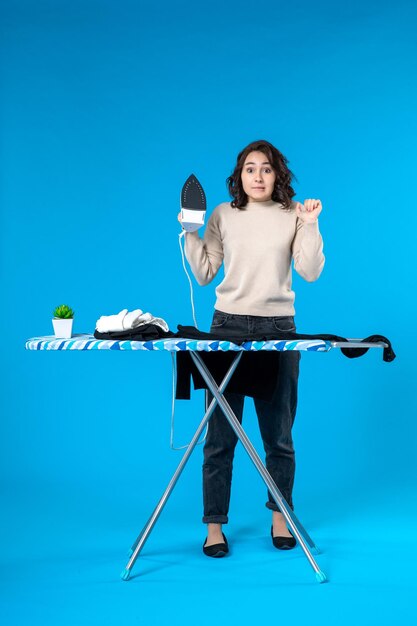 Front view of surprised young woman standing behind the board and holding iron on blue wave surface