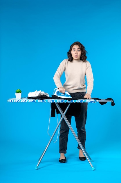 Front view of surprised young woman standing behind the board and ironing the cloth on blue wave background