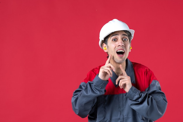 Free photo front view of surprised young worker in uniform with hard hat and wearing earplugs on isolated red wall