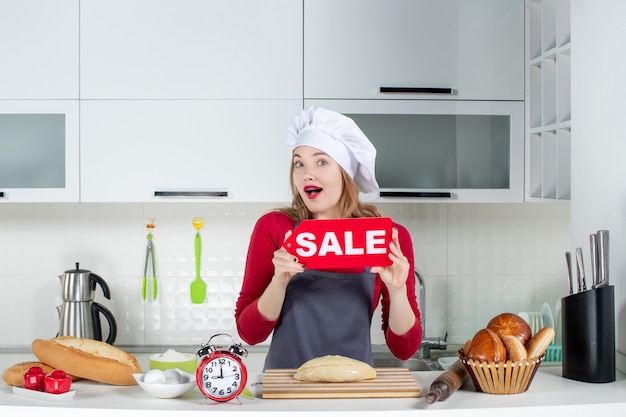 Front view sweet blonde woman in cook hat and apron holding up sale sign in the kitchen