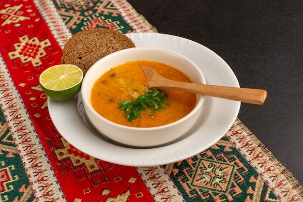 Zuppa di verdure gustosa vista frontale all'interno del piatto con pagnotta di pane e limone sullo scrittorio scuro.