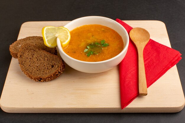 Zuppa di verdure gustosa vista frontale all'interno del piatto con fetta di limone e pagnotte di pane sulla scrivania scura.