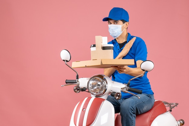 Front view of thankful male delivery person in mask wearing hat sitting on scooter showing orders on pastel peach background