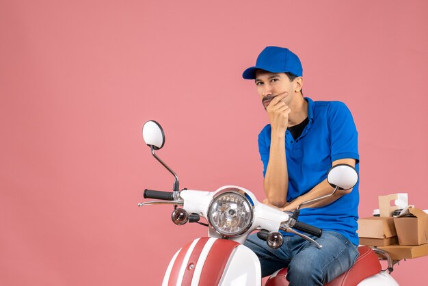 Front view of thinking delivery guy wearing hat sitting on scooter on pastel peach background