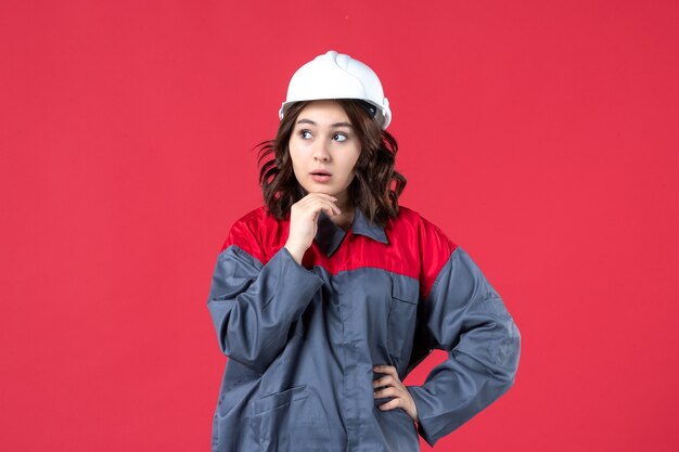 Вид спереди думающей женщины-строителя в униформе с каской на изолированном красном фоне