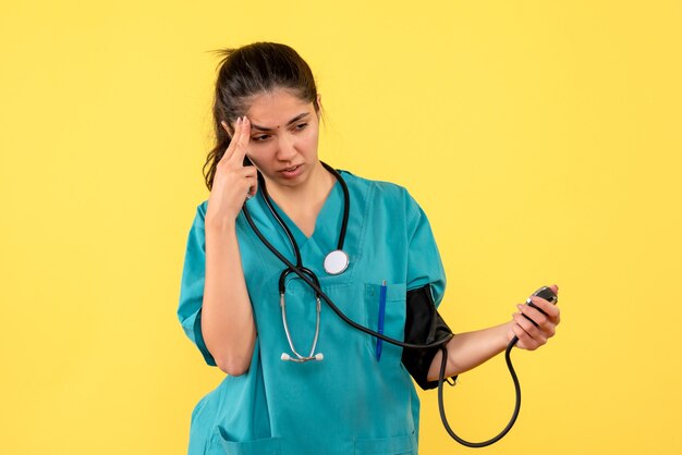 Front view thinking female doctor in uniform holding sphygmomanometers standing on yellow background