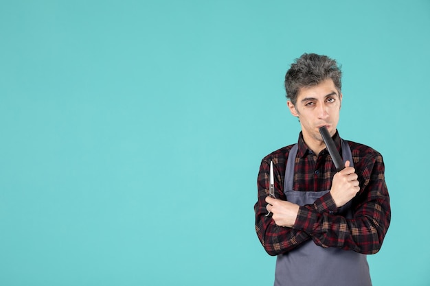 Front view of thinking male barber wearing gray apron and holding comb scissor on blue color surface