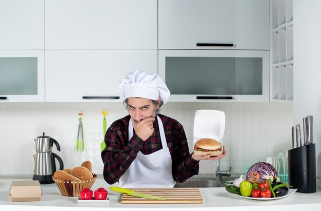 Front view thinking male chef holding up burger in modern kitchen