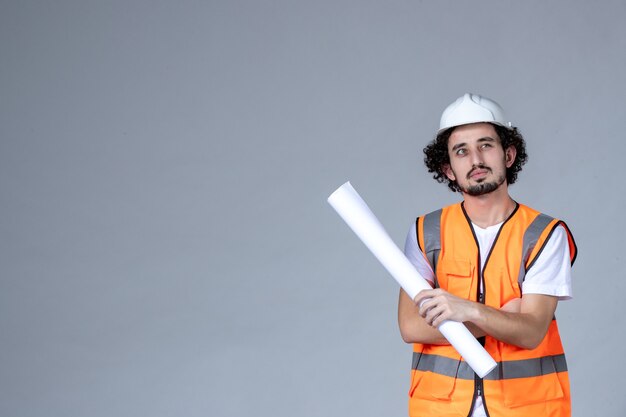 Front view of thinking male constructor in warning vest wearing safety helmet and holding blank on gray wave wall