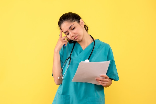 Front view thinking pretty female doctor holding papers on yellow background