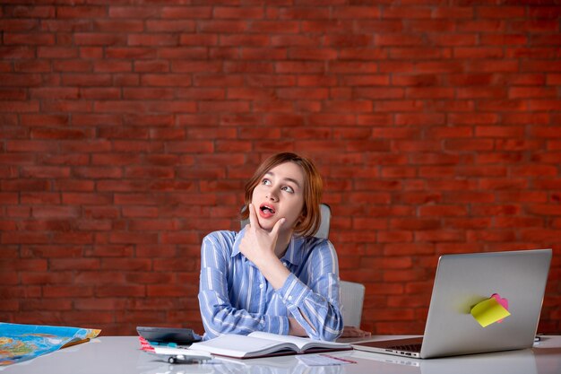 Front view thinking travel agent sitting behind her working place with laptop map agency service assistant ticket manager operator global