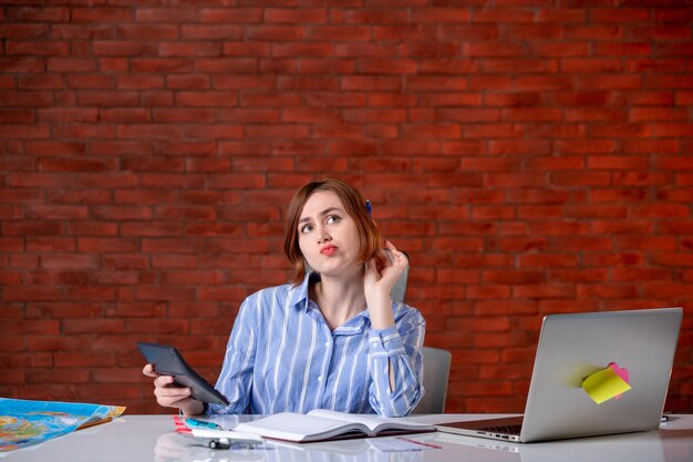 Front view thinking travel agent sitting behind her working place with laptop working with calculator map agency service assistant manager ticket operator world global