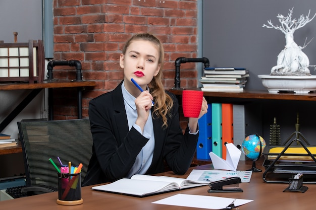 Front view of thinking young female sitting at a table and holding a red cup in the office