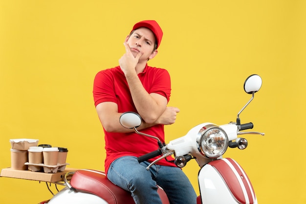 Front view of thinking young guy wearing red blouse and hat delivering orders on yellow background