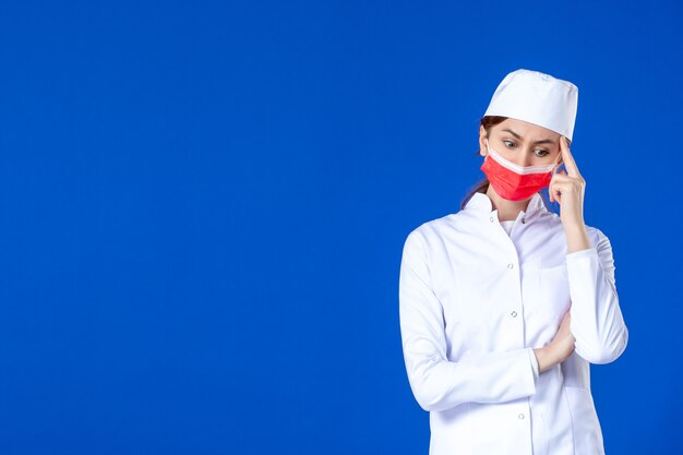 Front view thinking young nurse in medical suit with red protective mask on blue wall