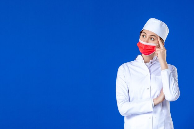 Front view thinking young nurse in medical suit with red protective mask on blue wall