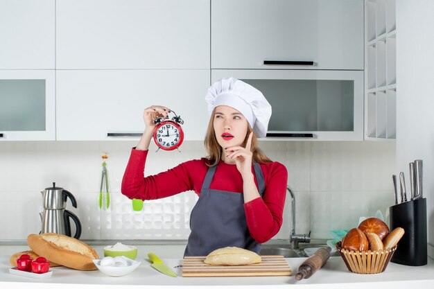 Front view thinking young woman holding red alarm clock in the kitchen