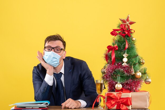 Front view of thoughtful business man sitting at the table near xmas tree and presents on yellow