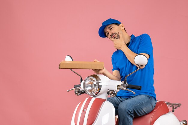 Front view of thoughtful courier man wearing hat sitting on scooter holding order on pastel peach background