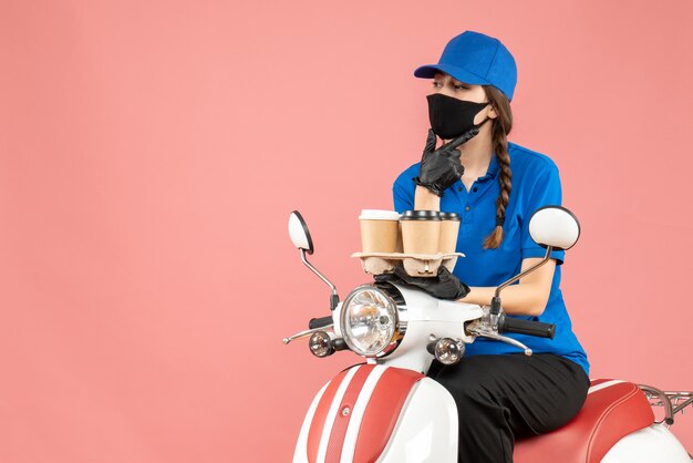 Вид спереди вдумчивого доставщика женского пола в медицинской маске и перчатках, сидящего на скутере и держащего заказы на пастельно-персиковом фоне