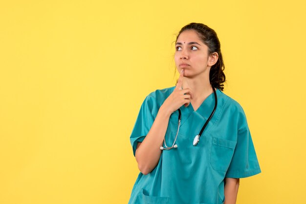 Front view thoughtful female doctor with stethoscope putting hand to her chin on yellow background