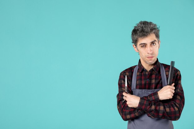 Front view of thoughtful male barber wearing gray apron and holding comb scissor on blue color background