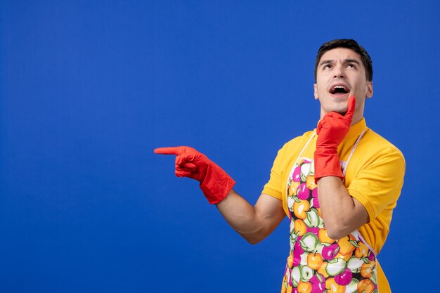 Front view of thoughtful male housekeeper in apron pointing at left standing on blue wall