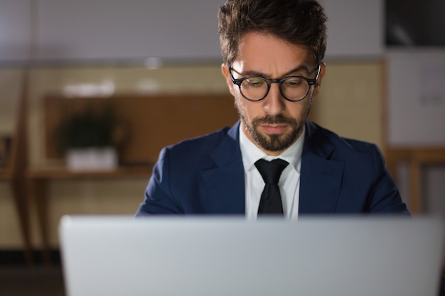 Front view of thoughtful man in eyeglasses typing at laptop