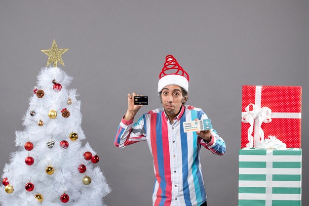 Front view thoughtful man holding card and travel ticket around xmas tree and presents