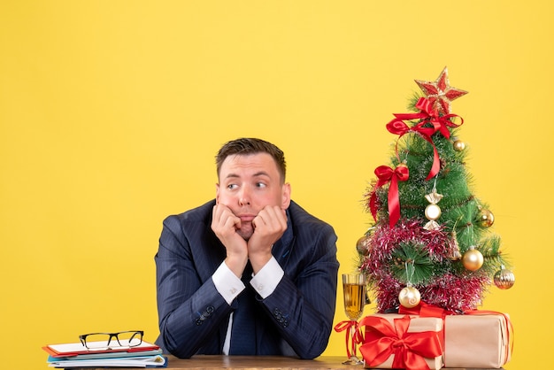 Vista frontale dell'uomo premuroso che si siede al tavolo vicino all'albero di natale e regali su giallo