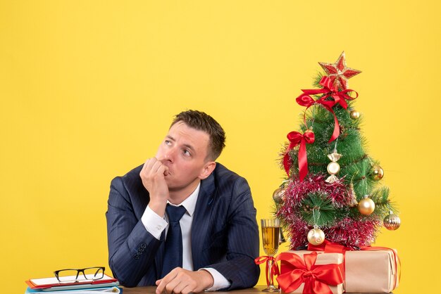 クリスマスツリーと黄色の贈り物の近くのテーブルに座っている何かについて考えている思いやりのある男の正面図