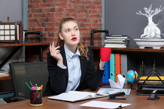 Free photo front view of thoughtful young female sitting at a table and holding a red cup in the office