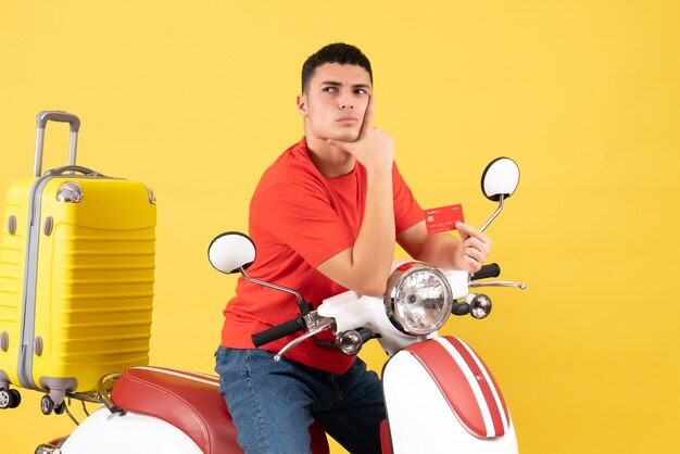 Front view thoughtful young man on moped holding discount card putting hand on his chin