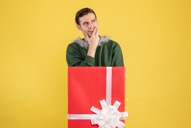 Front view thoughtful young man standing behind big giftbox on yellow 