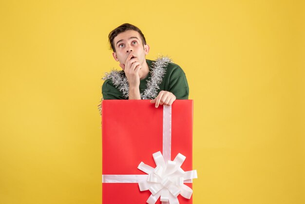 Front view thoughtful young man standing behind big giftbox on yellow 