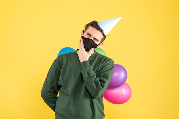 Front view thoughtful young man with party cap and colorful balloons standing on yellow 