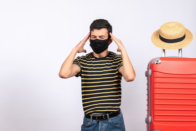 Vista frontale premuroso giovane turista con maschera nera in piedi vicino a valigia rossa tenendo la testa tra le mani