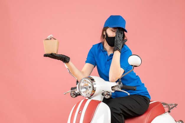 Front view of tired delivery person wearing medical mask and gloves sitting on scooter delivering orders suffering from headache on pastel peach background