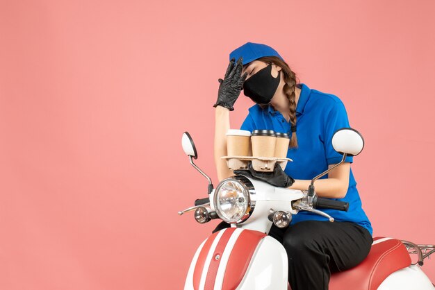 Вид спереди усталого доставщика женского пола в медицинской маске и перчатках, сидящего на скутере и держащего заказы на пастельном персиковом фоне