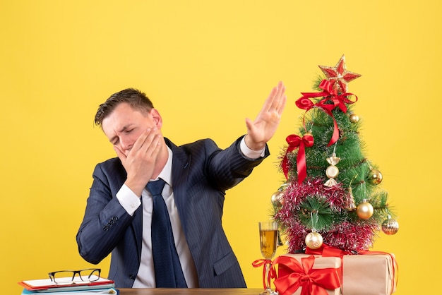クリスマスツリーと黄色の贈り物の近くのテーブルに座ってあくびをしている疲れた男の正面図