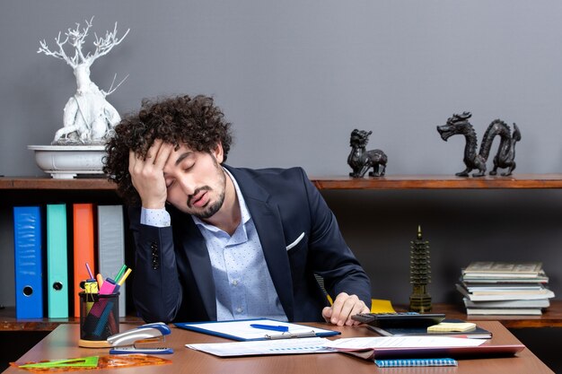 Front view of tired young businessman sitting at desk in office