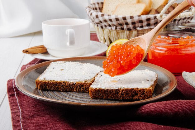 Front view toasts with butter and a spoon of red caviar on a plate with a cup of tea