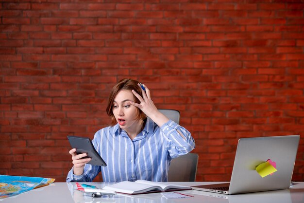 Front view travel agent sitting behind her working place with laptop and notepad indoors map agency service managers assistant ticket world global