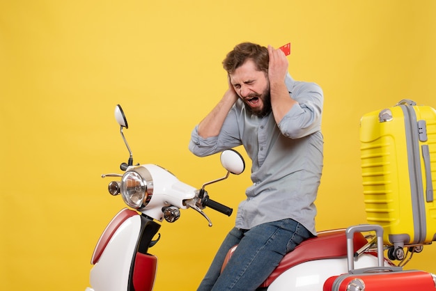 Front view of travel concept with angry nervous emotional young man sitting on motocycle with suitcases on it holding bank card on yellow 